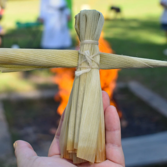 A hand holds a corn dolly, a bonfire in the background